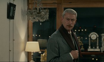 Movie still from “Muriel” (1963), directed by Alain Resnais – A man in a suit and tie standing in front of a lamp; Medium shot, Over the shoulder angle