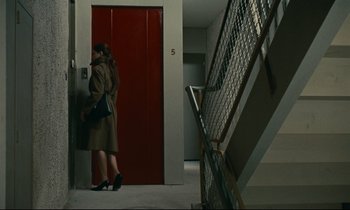 Movie still from “Muriel” (1963), directed by Alain Resnais – A woman standing in front of a red door next to a staircase; Wide shot, High angle