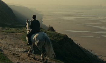 Movie still from “Muriel” (1963), directed by Alain Resnais – A man riding a horse down a hill next to a body of water; Extreme Wide shot, High angle