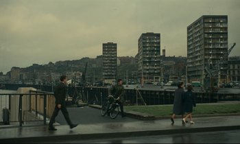 Movie still from “Muriel” (1963), directed by Alain Resnais – A group of people walking on a sidewalk near a city; Extreme Wide shot, High angle