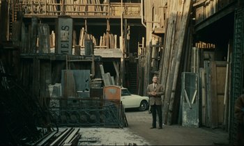 Movie still from “Muriel” (1963), directed by Alain Resnais – A man in a suit standing next to a car; Wide shot, High angle