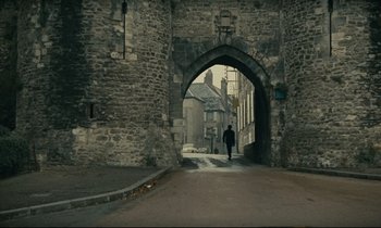 Movie still from “Muriel” (1963), directed by Alain Resnais – A person walking down a street under an archway; Extreme Wide shot, Over the shoulder angle