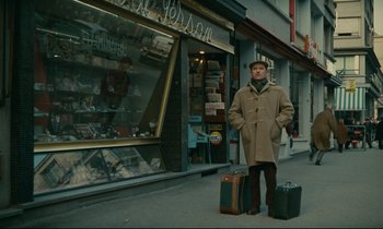Movie still from “Muriel” (1963), directed by Alain Resnais – A man standing on the side of the street with luggage; Wide shot, High angle
