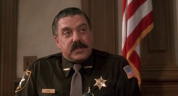 Movie still from “My Cousin Vinny” (1992), directed by Jonathan Lynn – A man in a police uniform sitting in front of an american flag; Close Up shot, Low angle