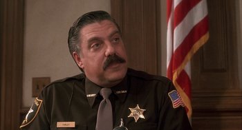 Movie still from “My Cousin Vinny” (1992), directed by Jonathan Lynn – A man in a sheriff's uniform sitting in front of an american flag; Close Up shot, Low angle