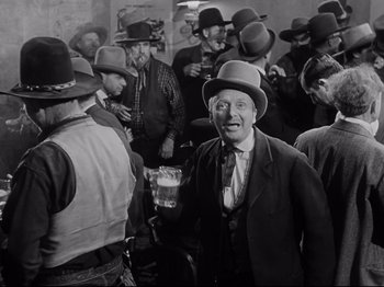 Movie still from “My Darling Clementine” (1946), directed by John Ford – An old man in a suit and hat is holding a glass of beer in front of a group of men; Medium shot, Low angle