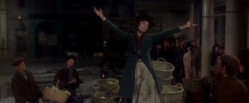 Movie still from “My Fair Lady” (1964), directed by George Cukor – A woman wearing a hat and holding her arms in the air; Medium shot, Low angle