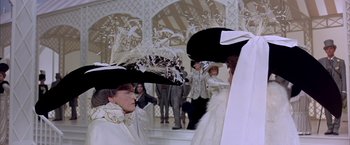 Movie still from “My Fair Lady” (1964), directed by George Cukor – Two women wearing fancy hats in front of a group of people; Medium shot, Over the shoulder angle
