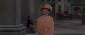 Movie still from “My Fair Lady” (1964), directed by George Cukor – A woman wearing a hat standing in front of a building; Medium shot, Over the shoulder angle