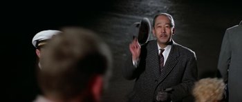 Movie still from “My Geisha” (1962), directed by Jack Cardiff – A man wearing a suit and a hat is holding his hand up; Medium shot, Over the shoulder angle