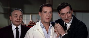 Movie still from “My Geisha” (1962), directed by Jack Cardiff – A man in a lab coat is looking at the camera while another man adjusts his tie; Close Up shot, Over the shoulder angle