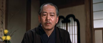 Movie still from “My Geisha” (1962), directed by Jack Cardiff – An older man wearing a black robe and a black hat; Close Up shot, Low angle