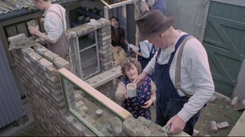 Movie still from “My Left Foot” (1989), directed by Jim Sheridan – A woman holding a cup in her hand while standing in front of a brick wall; Wide shot, High angle