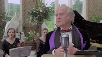 Movie still from “My Left Foot” (1989), directed by Jim Sheridan – An older man wearing a black jacket and a purple tie; Medium shot, Over the shoulder angle