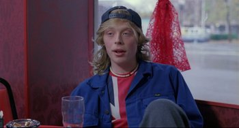 Movie still from “My Own Private Idaho” (1991), directed by Gus Van Sant – A man with long blonde hair sitting at a table with a glass; Medium shot, Over the shoulder angle