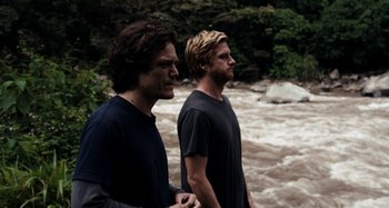Movie still from “My Son, My Son, What Have Ye Done” (2009), directed by Werner Herzog – Two young men standing in front of a rushing river side by side; Medium shot, Over the shoulder angle
