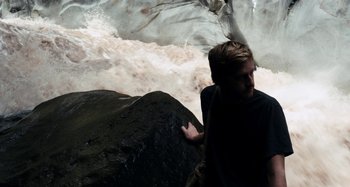 Movie still from “My Son, My Son, What Have Ye Done” (2009), directed by Werner Herzog – A man standing next to a large rock in the water; Medium shot, High angle