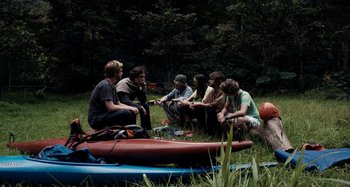 Movie still from “My Son, My Son, What Have Ye Done” (2009), directed by Werner Herzog – A group of people sitting in the grass next to a canoe; Wide shot, High angle