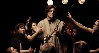 Movie still from “My Son, My Son, What Have Ye Done” (2009), directed by Werner Herzog – A group of people standing around a man holding a sword; Medium shot, Over the shoulder angle