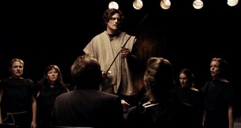 Movie still from “My Son, My Son, What Have Ye Done” (2009), directed by Werner Herzog – A man holding a sword in front of a group of people; Medium shot, Over the shoulder angle