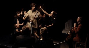 Movie still from “My Son, My Son, What Have Ye Done” (2009), directed by Werner Herzog – A group of people in a room playing musical instruments; Wide shot, Over the shoulder angle