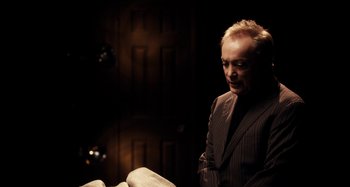 Movie still from “My Son, My Son, What Have Ye Done” (2009), directed by Werner Herzog – A man in a suit standing in front of a book; Medium shot, High angle