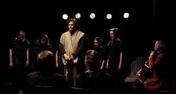 Movie still from “My Son, My Son, What Have Ye Done” (2009), directed by Werner Herzog – A group of people standing in front of a man holding a sword; Wide shot, Over the shoulder angle