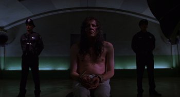 Movie still from “Mystery Men” (1999), directed by Kinka Usher – A man with long hair is sitting in a dark room; Medium shot, Low angle