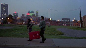 Movie still from “Mystery Train” (1989), directed by Jim Jarmusch – A man and a woman walking down a sidewalk with luggage; Wide shot, Over the shoulder angle