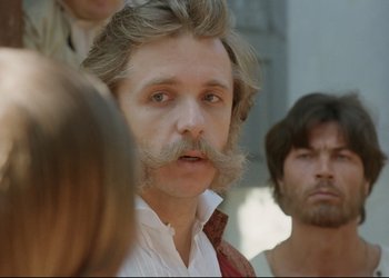 Movie still from “A Few Days from the Life of I.I. Oblomov” (1980), directed by Nikita Mikhalkov – A man with long hair and a mustache is standing in front of a group of people; Close Up shot, Over the shoulder angle