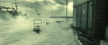 Movie still from “Oceans” (2009), directed by Jacques Cluzaud – A large wave crashes into a pier and a beach chair; Extreme Wide shot, High angle