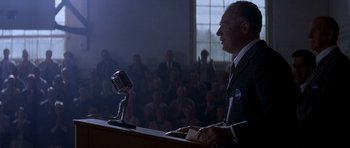 Movie still from “October Sky” (1999), directed by Joe Johnston – A man is speaking at a microphone in front of a crowd; Medium shot, Low angle