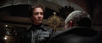 Movie still from “Octopussy” (1983), directed by John Glen – A man in a leather jacket is talking to another man in front of a cross; Close Up shot, Low angle