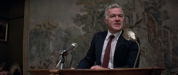 Movie still from “Octopussy” (1983), directed by John Glen – A man wearing a suit and tie sitting in front of a microphone; Close Up shot, High angle