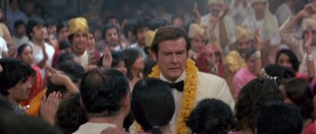 Movie still from “Octopussy” (1983), directed by John Glen – A man in a white suit with a yellow lei; Medium shot, Over the shoulder angle