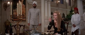 Movie still from “Octopussy” (1983), directed by John Glen – A woman sitting at a dinner table with a man standing behind her; Medium shot, Low angle