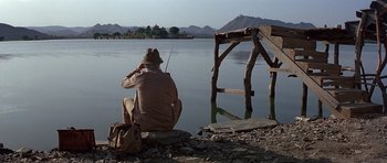 Movie still from “Octopussy” (1983), directed by John Glen – A man sitting on the shore of a body of water fishing; Wide shot, Low angle