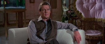 Movie still from “Octopussy” (1983), directed by John Glen – A man sitting on top of a couch in a living room; Medium shot, Over the shoulder angle