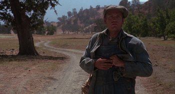 Movie still from “Of Mice and Men” (1992), directed by Gary Sinise – A man standing on the side of a dirt road holding a cell phone; Medium shot, High angle