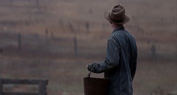 Movie still from “Of Mice and Men” (1992), directed by Gary Sinise – A man in a hat holding a brown bag in a field; Wide shot, Over the shoulder angle