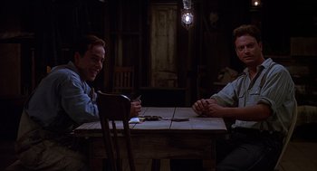 Movie still from “Of Mice and Men” (1992), directed by Gary Sinise – Two men sitting at a wooden table in a dark room; Medium shot, Low angle