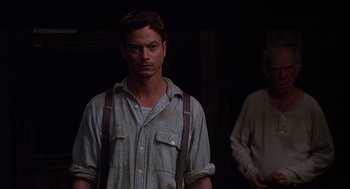 Movie still from “Of Mice and Men” (1992), directed by Gary Sinise – A man standing in a dark room wearing suspenders; Close Up shot, Over the shoulder angle