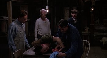 Movie still from “Of Mice and Men” (1992), directed by Gary Sinise – A group of men standing around a table with a man laying on top of it; Medium shot, High angle