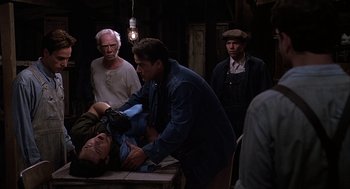 Movie still from “Of Mice and Men” (1992), directed by Gary Sinise – A group of men standing around a man on a table; Medium shot, Over the shoulder angle