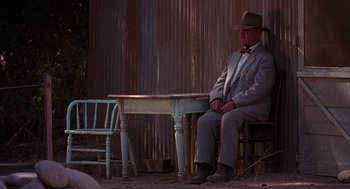 Movie still from “Of Mice and Men” (1992), directed by Gary Sinise – A man sitting at a table in front of a wooden fence; Wide shot, Low angle