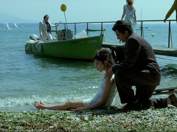 Movie still from “Oh, Woe Is Me” (1993), directed by Jean-Luc Godard – A man and a woman sitting on the shore of a body of water; Wide shot, Low angle