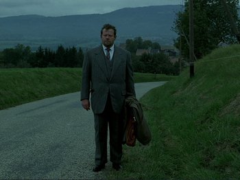 Movie still from “Oh, Woe Is Me” (1993), directed by Jean-Luc Godard – A man in a suit and tie standing on the side of a road; Wide shot, Low angle