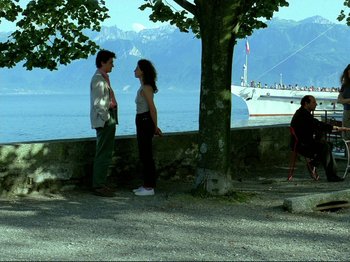 Movie still from “Oh, Woe Is Me” (1993), directed by Jean-Luc Godard – A man and a woman standing next to each other near the water; Wide shot, High angle