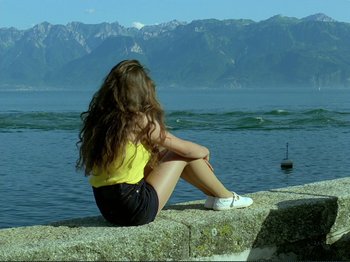 Movie still from “Oh, Woe Is Me” (1993), directed by Jean-Luc Godard – A woman sitting on the edge of a pier looking out at the ocean; Wide shot, High angle