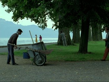 Movie still from “Oh, Woe Is Me” (1993), directed by Jean-Luc Godard – A man pushing a wheelbarrow full of trash on the side of the road; Extreme Wide shot, Over the shoulder angle